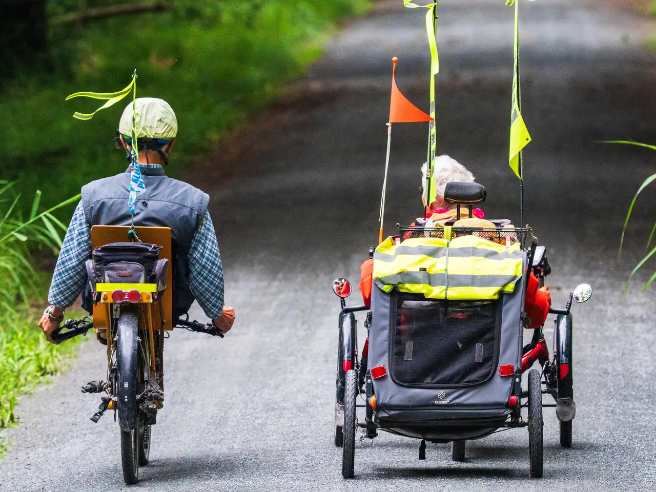 Trailer with safety flags and reflectors Two recumbent riders seen from behind on a paved path, one towing a covered two-wheel trailer fitted with safety flags, reflectors and a hi-vis cover