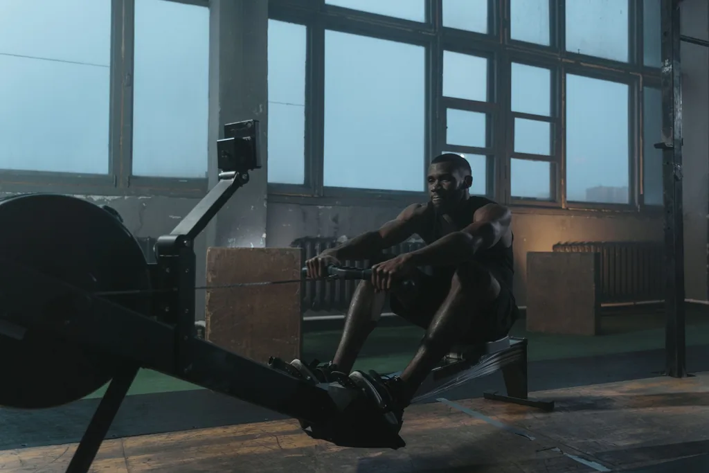Rowing: moderate aerobic transfer, high upper body and core work, low injury risk Athlete at full stretch on an indoor rowing machine in a high-windowed gym, the whole machine and rower in frame