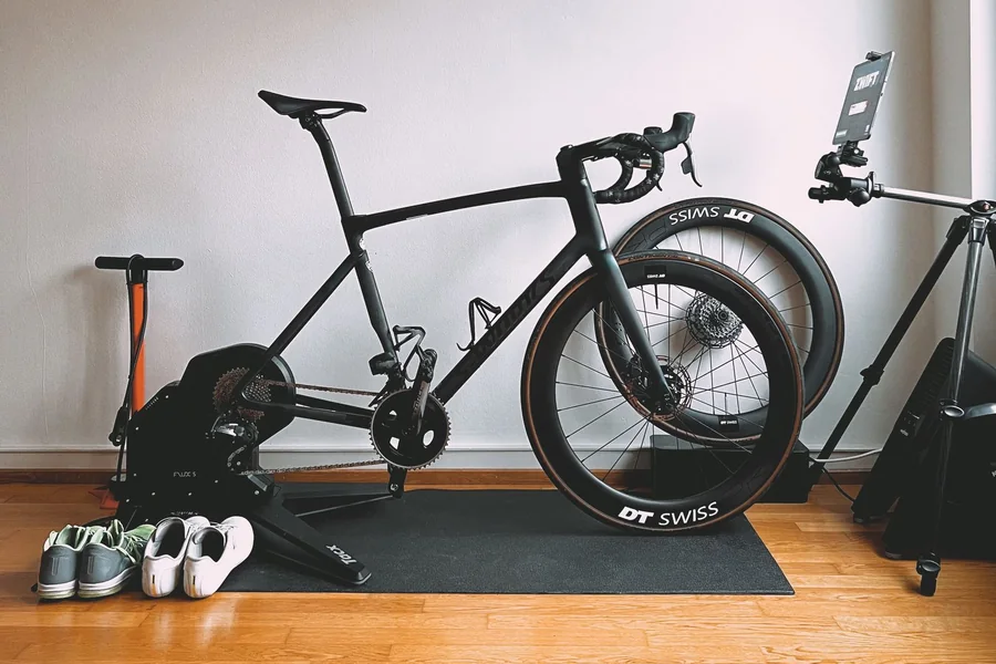 Road bike mounted on a direct-drive smart trainer on a mat, with a tablet running Zwift, shoes and a floor pump alongside