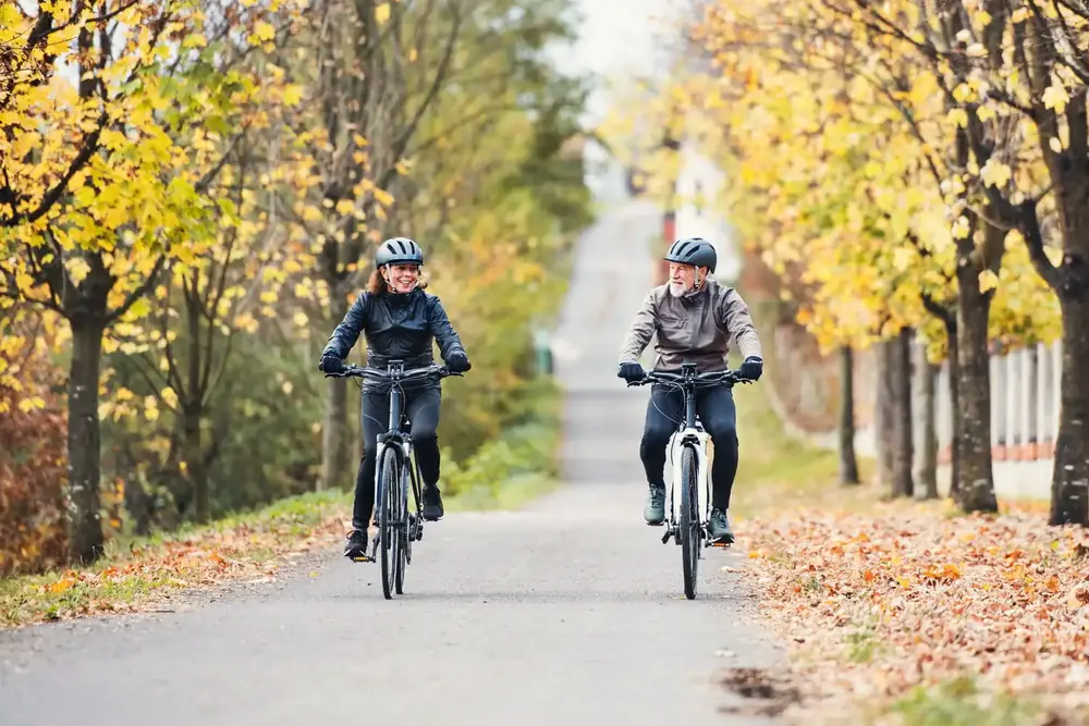 Riding e-bikes in cool autumn weather Two riders in jackets and gloves riding e-bikes side by side down a tree-lined road covered in fallen autumn leaves
