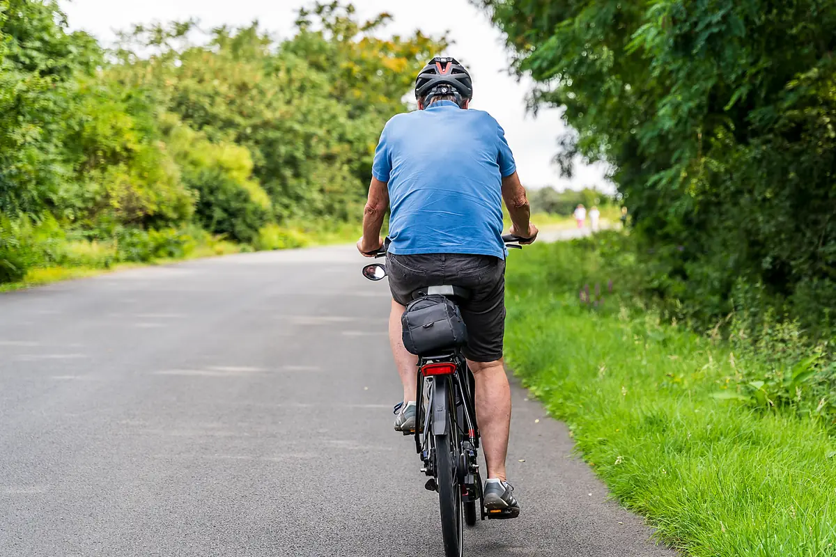 Older cyclist seen from behind riding a quiet tree-lined road, keeping the engine going with age