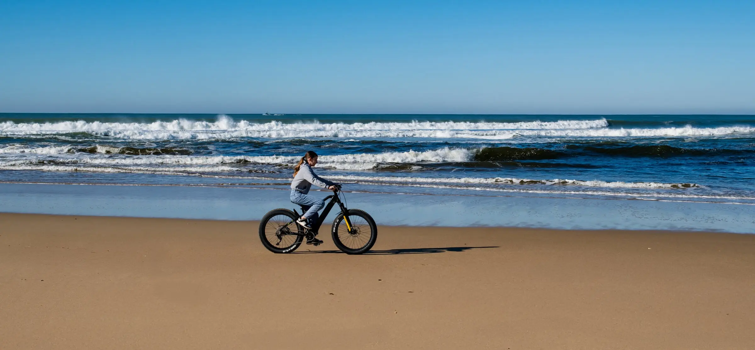 Rider on a black fat-tire e-bike along the shoreline with waves behind Rider on a black fat-tire e-bike along the shoreline with waves behind