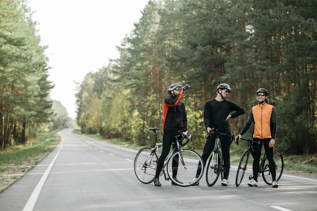 Three road cyclists in helmets standing with their bikes on a quiet forest road during a long ride
