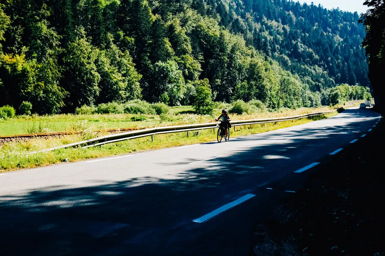 Route selection is the road cyclist's single biggest safety lever Cyclist riding along a quiet two-lane road through a forested valley with a car far in the distance