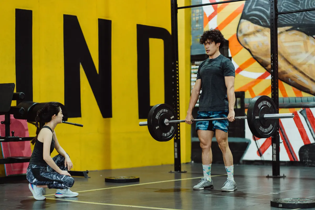 Man deadlifting a barbell in a gym while a trainer crouches at the side watching the speed of the bar Man deadlifting a barbell in a gym while a trainer crouches at the side watching the speed of the bar