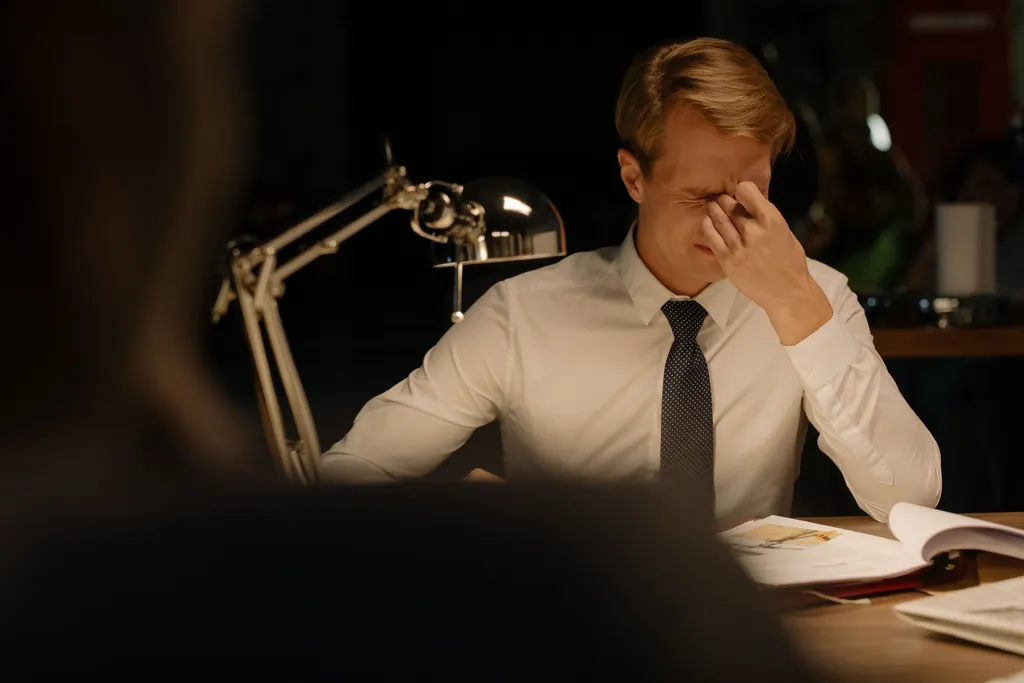 Man in a shirt and tie at a desk pinching the bridge of his nose over an open file Man in a shirt and tie at a desk pinching the bridge of his nose over an open file